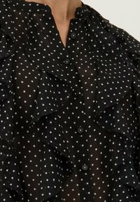 Zwarte transparante blouse met witte stippen, met ruches aan de voorkant, een kraag en zwarte knopen.