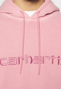 Růžová mikina s kapucí z měkké látky s výšivkou loga "carhartt" v mírně tmavší růžové barvě. Má nastavitelný stahovací šňůrku na kapuci.
