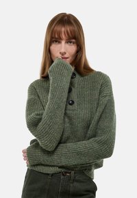 Pull en maille verte avec un col montant, doté de trois boutons noirs, de poignets côtelés et d'une finition texturée, associé à un pantalon sombre.