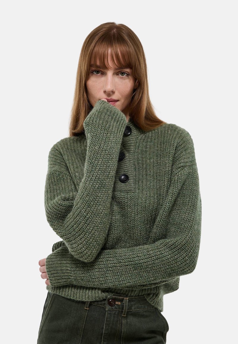 Pull en maille verte avec un col montant, doté de trois boutons noirs, de poignets côtelés et d'une finition texturée, associé à un pantalon sombre.