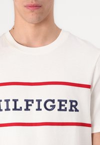 White cotton T-shirt with a round neckline, featuring a red horizontal stripe and navy blue "HILFIGER" text embroidered in the center.