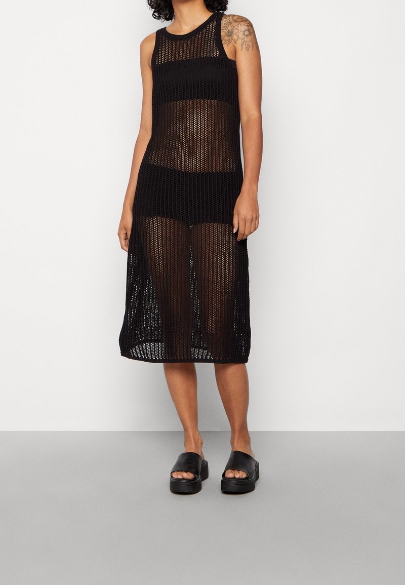 Abito lungo in maglia nera con design senza maniche, texture a rete aperta e lunghezza fino al polpaccio. Abbinato a sandali con zeppa neri.