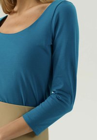 Donna che indossa un top a maniche lunghe teal con scollatura a barca e una gonna beige, tagliata all'altezza del petto e del braccio.