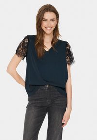 Top blu navy con scollatura a V e maniche corte in pizzo nero. Tessuto morbido con un design leggermente drappato, indossato con jeans scuri.
