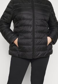 Giacca a vento nera con cerniera frontale, motivo trapuntato orizzontale e tasche laterali. Materiale morbido, leggero con una texture liscia.