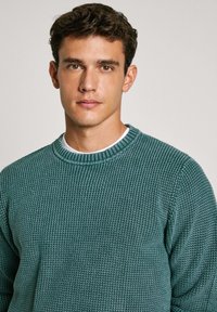 Teal pulover iz teksturirane pletenine z okroglim izrezom in rebrastimi manšeti. Obut nad belim srajcem, ki ima subtilen tkan vzorec.