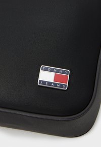 Svart teksturert materiale pouch med et metall Tommy Jeans-logo med blå, hvite og røde fargeblokker. Glatte kanter og subtile detaljer.