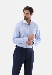 Helles blaues Hemd mit weißem Kragen, langärmlig und körpernah geschnitten. Kombiniert mit dunkelblauen Hosen.