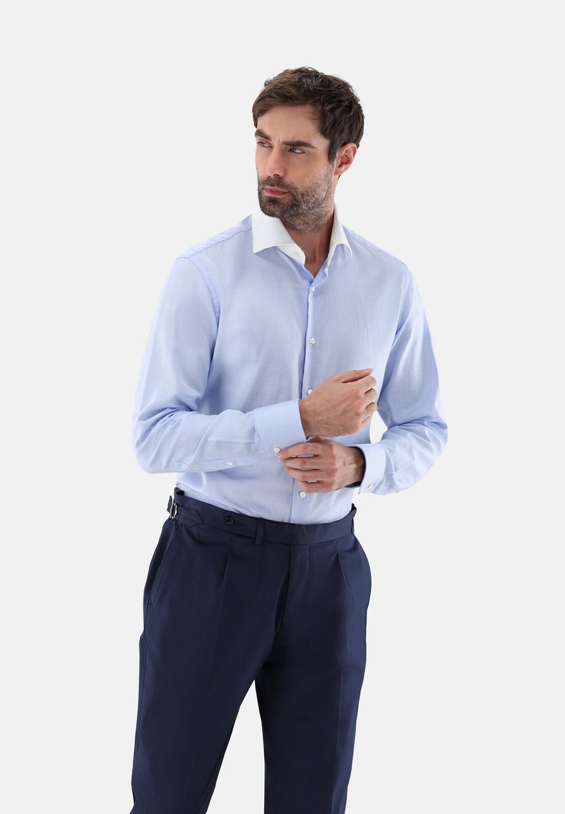 Helles blaues Hemd mit weißem Kragen, langärmlig und körpernah geschnitten. Kombiniert mit dunkelblauen Hosen.