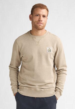 Roodharige man met lichte gezichtsbeharing die een beige trui met een bloempatch draagt, handen in de zakken, tegen een effen witte achtergrond.