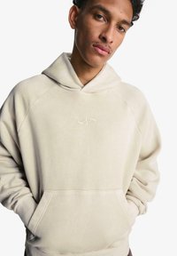 Beige mikina s kapucí vyrobená z měkké tkaniny, se přední kapsou a jemným vyšitým logem na hrudi, s raglánovými rukávy.