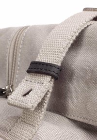 Dettaglio di una borsa in tela beige con un accento di tracolla in pelle nera, hardware argentato e una superficie testurizzata con cuciture visibili.