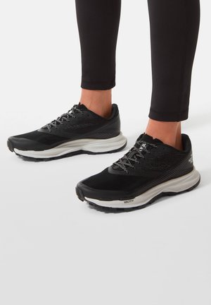 Osoba w czarno-białych sportowych butach do biegania z wzorzystymi sznurówkami i czarnych legginsach, stojąca na jasnoszarej powierzchni.