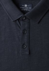 Mörkblå pikétröja i strukturerad tyg, med klassisk krage, tre knappar och en lapp med varumärkesnamn och designinformation.