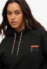 Hoodie noir avec poche kangourou, marque rouge et cordons texturés. Présente des embouts en métal argenté et des détails imprimés sur le côté.