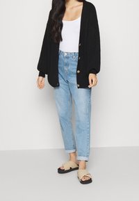 Kvinna iklädd en svart oversized kofta, vit tanktopp, ljusblå uppvikta jeans och beige sandaler med korsband som står på ett grått golv.