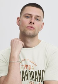 Svetlo siva majica s grafičkim printom u tamnozelenim i narančastim tonovima. Kratki rukavi, okrugli izrez, i srebrna narukvica na zapešću.