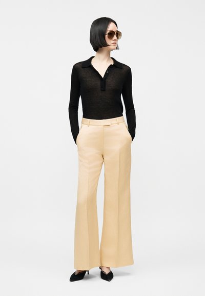 Kvinne med kort svart hår iført store solbriller, svart langermet topp, beige vidbukser og svarte spisse hæler, stående med hendene i lommene.