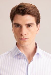 Jonge man met kort bruin haar, gekleed in een lichtblauw gestreept overhemd met knopen, die rechtop kijkt met een neutrale uitdrukking.