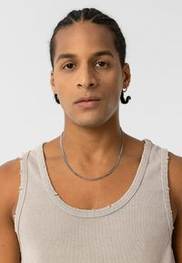 Grijze geribbelde tanktop met rafelige randen en een zilveren kettinghalsketting gedragen door een persoon met vlechten, neutrale achtergrond.