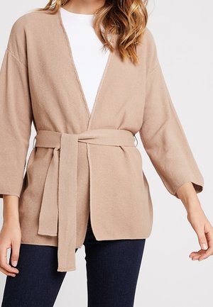 Beiger Cardigan mit Gürtel, getragen über einem weißen Oberteil und kombiniert mit dunkelblauen Hosen, gezeigt an einer Person von den Schultern bis zur Hüfte.