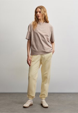 Kvinde står iført en taupe oversized t-shirt, cremefarvede joggingbukser og hvide sneakers mod en ensfarvet lysegrå baggrund.