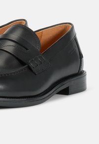 Svarta läderloafers med en slät yta, som har ett dekorativt band över framfoten och en låg rektangulär klack. Inuti är de i tan-färg.