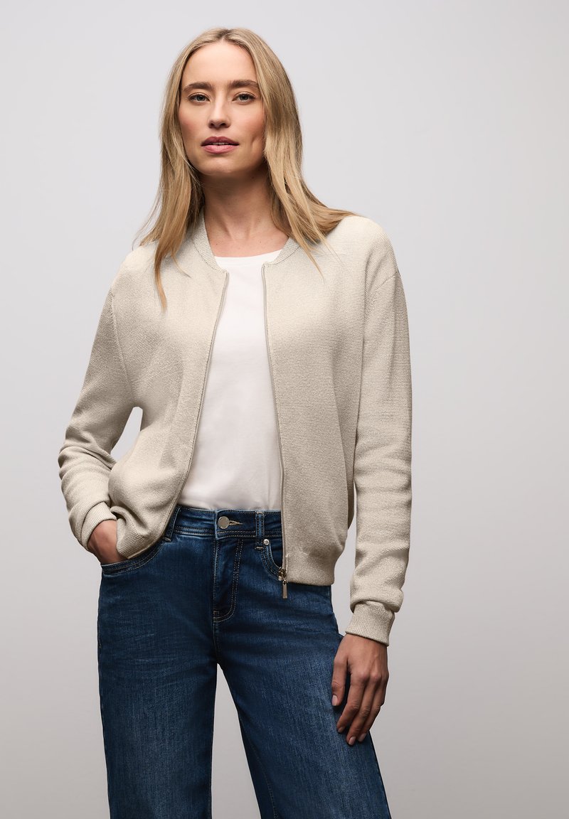 Junge Frau mit langem blondem Haar, die einen beigen Pullover mit Reißverschluss über einem weißen Hemd und blauen Jeans trägt, vor einem schlichten hellen Hintergrund stehend.