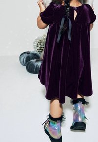 Vestido de terciopelo morado con mangas abullonadas, que presenta un botón en la parte trasera y un lazo. Combinado con botas de tobillo iridiscentes adornadas con volantes negros.