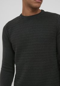 Temno siva pletena jopica z teksturiranim vzorcem, okroglo izrezana in z dolgimi rokavi z rebrastimi manšetami. Material se zdi mehak in topel.
