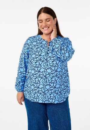 Camicia azzurra a maniche lunghe con un motivo floreale astratto in nero, scollatura rotonda e dettagli con bottoni, abbinata a jeans in denim blu.