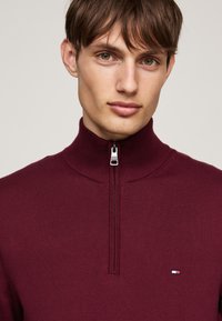 Burgundowa bluza zapinana na zamek z wysokim kołnierzem, o prążkowanej fakturze, z małą naszywką z logo w czerwonych, białych i niebieskich akcentach.