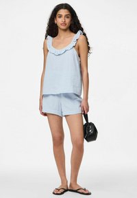 Gaiši zils bezpiedurkņu tops ar ruffles apkakli, apvienots ar atbilstošām šortiem. Modelis tur mazu melnu somu, valkājot melnus flip-flopus.