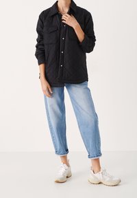 Svart vadderad skjortjacka med två bröstfickor, matchad med ljusblå jeans med uppvikta fållar och vita sneakers med beige detaljer.