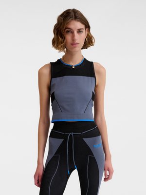 Kobieta w czarno-szarym topie na ramiączkach i dopasowanych legginsach z wysokim stanem z niebieskimi akcentami, stojąca na białym tle.