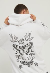 Hvit genser med grafisk design som har sommerfugler, to fugler, en rose, dekorative stjerner og teksten "DIN TUR" i svart blekk.