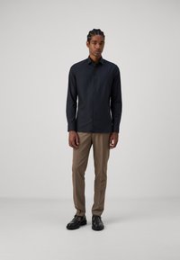 Schwarzes Hemd mit Knöpfen, slim fit, langärmlig und mit klassischem Kragen, kombiniert mit beigen maßgeschneiderten Hosen und schwarzen Schuhen.