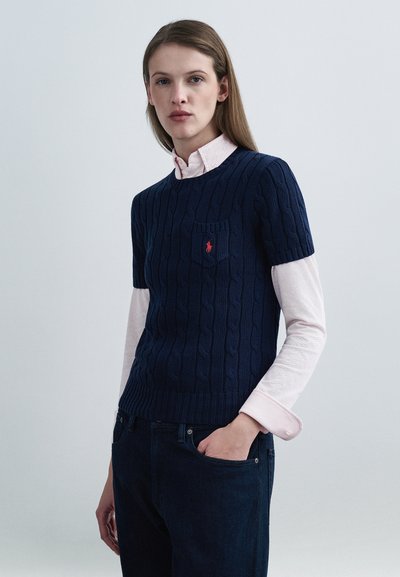 Pull à manches courtes en maille torsadée bleu marine avec une poche poitrine et un logo rouge, superposé à une chemise à col rose clair, associé à un jean foncé.