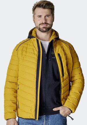 Gelbe Steppjacke mit horizontalem Steppmuster, Kapuze, vorderem Reißverschluss und Seitentaschen. Tragend über einem marineblauen Reißverschluss-Hoodie.