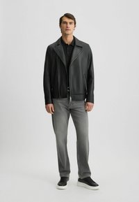 Giacca da biker in pelle nera con ampio colletto e zip frontale asimmetrica, abbinata a jeans grigi e sneakers nere con suola bianca.