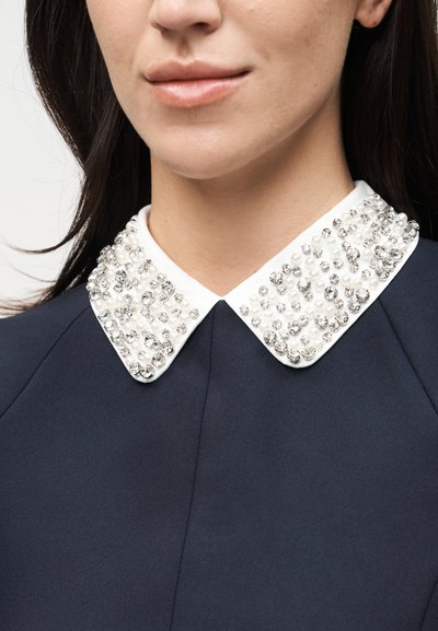 Γυναίκα που φοράει ναυτικό σύνολο με λευκό γιακά διακοσμημένο με κρύσταλλα και μαργαριτάρια, που δείχνει μέρος του κάτω μέρους του προσώπου και του λαιμού της.