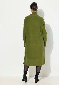 Robe tricotée verte longue avec un col haut et un ourlet côtelé, présentant une silhouette décontractée et des manches longues, associée à des bottes noires à la cheville.