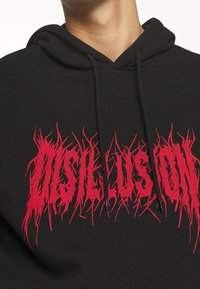 Černá mikina s kapucí, na které je výrazný zubatý červený potisk "DISHUSION" na hrudi. Materiál vypadá jemně a má texturovaný vzhled, k dispozici je také kapuce.