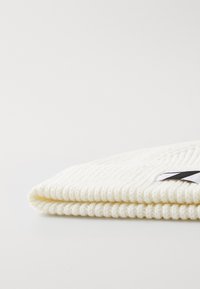 Bonnet en tricot couleur écrue, avec une texture côtelée, un revers et une étiquette noire et blanche cousue sur le côté.