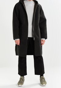 Musta, fleece-muotoinen pitkä takki, jossa on vetoketjullinen etuosa, huppu ja sivutaskut. Päällä vaaleanharmaa paita ja tummat manseryyrihousut vihreiden tennareiden kanssa.