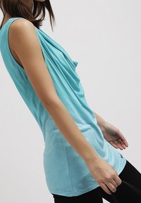 Un top senza maniche di un turchese chiaro con un design del collo drappeggiato, texture liscia e una vestibilità rilassata, abbinato a leggings neri.