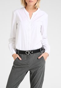 Valge krae blouse, millel on eesmine nööpide rida, koos hallide ülikonnatrekkidega ja musta vööd, millel on hõbedane CK-logo.