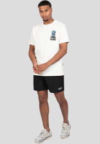 Camiseta de algodón crema con estampado azul y texto negro, combinada con pantalones cortos negros. Zapatillas grises visibles, de pie sobre un fondo liso.