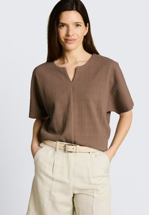 Frau mit langen dunklen Haaren trägt ein strukturiertes braunes Kurzarmshirt und hoch taillierte beige Hose mit beigem Gürtel, Hände in den Taschen.