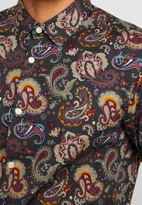 Lyhythihainen paita tummasta kankaasta, jossa monimutkaiset paisley-kuviot punaisena, vihreänä ja keltaisena, valkoisilla napin koristeilla ja klassisella kauluksella.